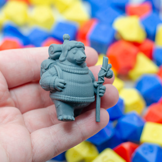 Gray 3d printed figurine of a bear in a sweater holding a staff, surrounded by colorful polyhedral dice, held in a hand to show scale.