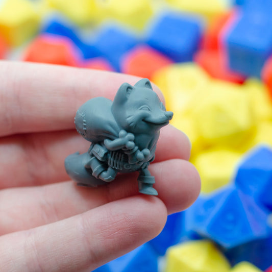 Gray figurine of a fox thief with a sneaky expression, held in a hand with colorful dice in the background