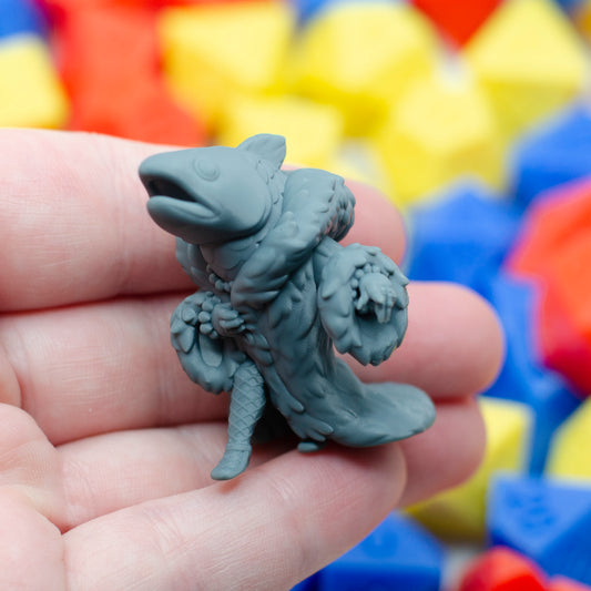 Gray 3d printed miniature figurine of a reverse mermaid held in a hand with colorful dice in the background