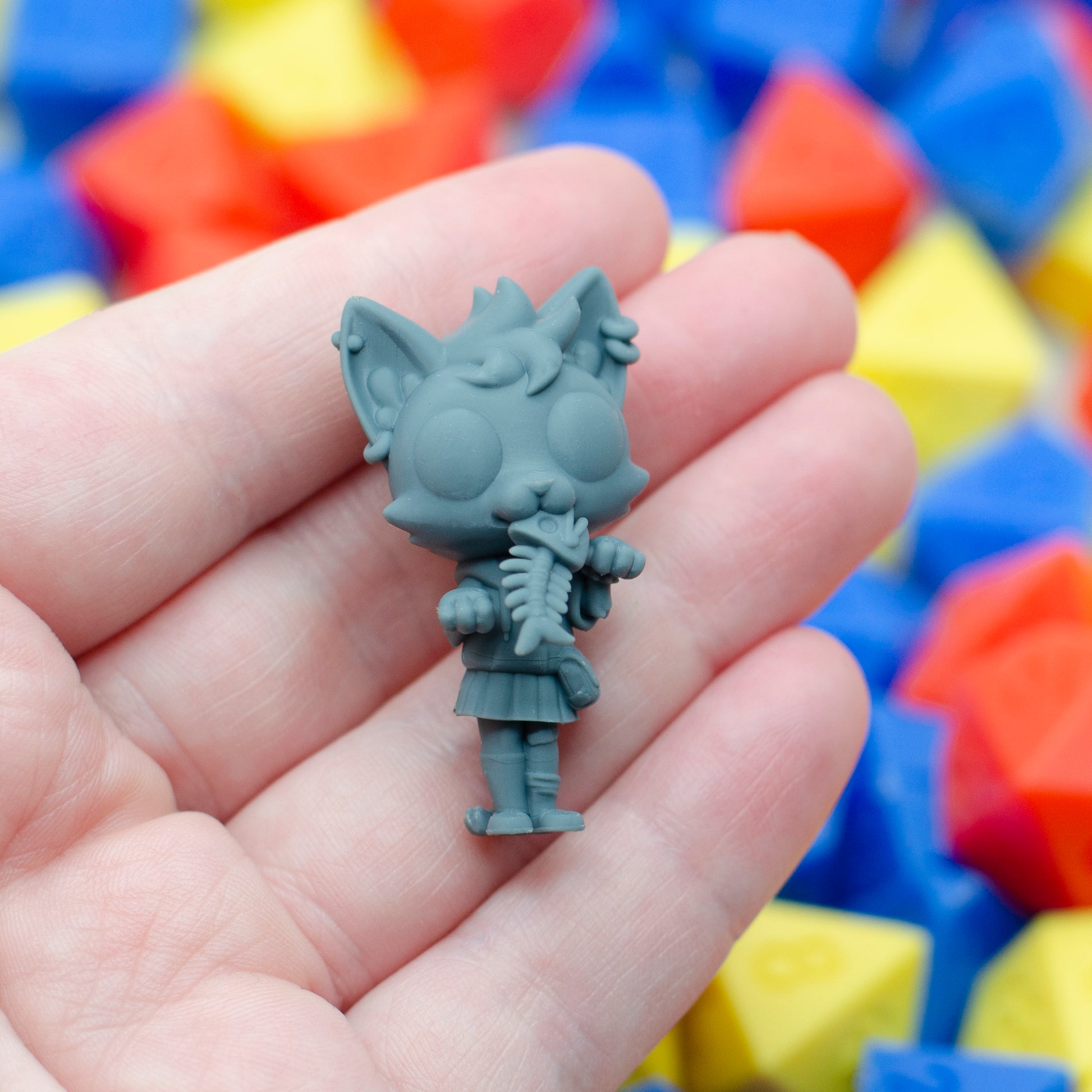 Gray 3d printed TTRPG miniature figurine of a catgirl with a school uniform and a fish bones in her mouth. It is held in a hand with colorful geometric blocks in the background