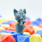 Gray resin printed 3d miniature of a cat girl standing in a trash can, with fish bones in her mouth. She stands on colorful dice.