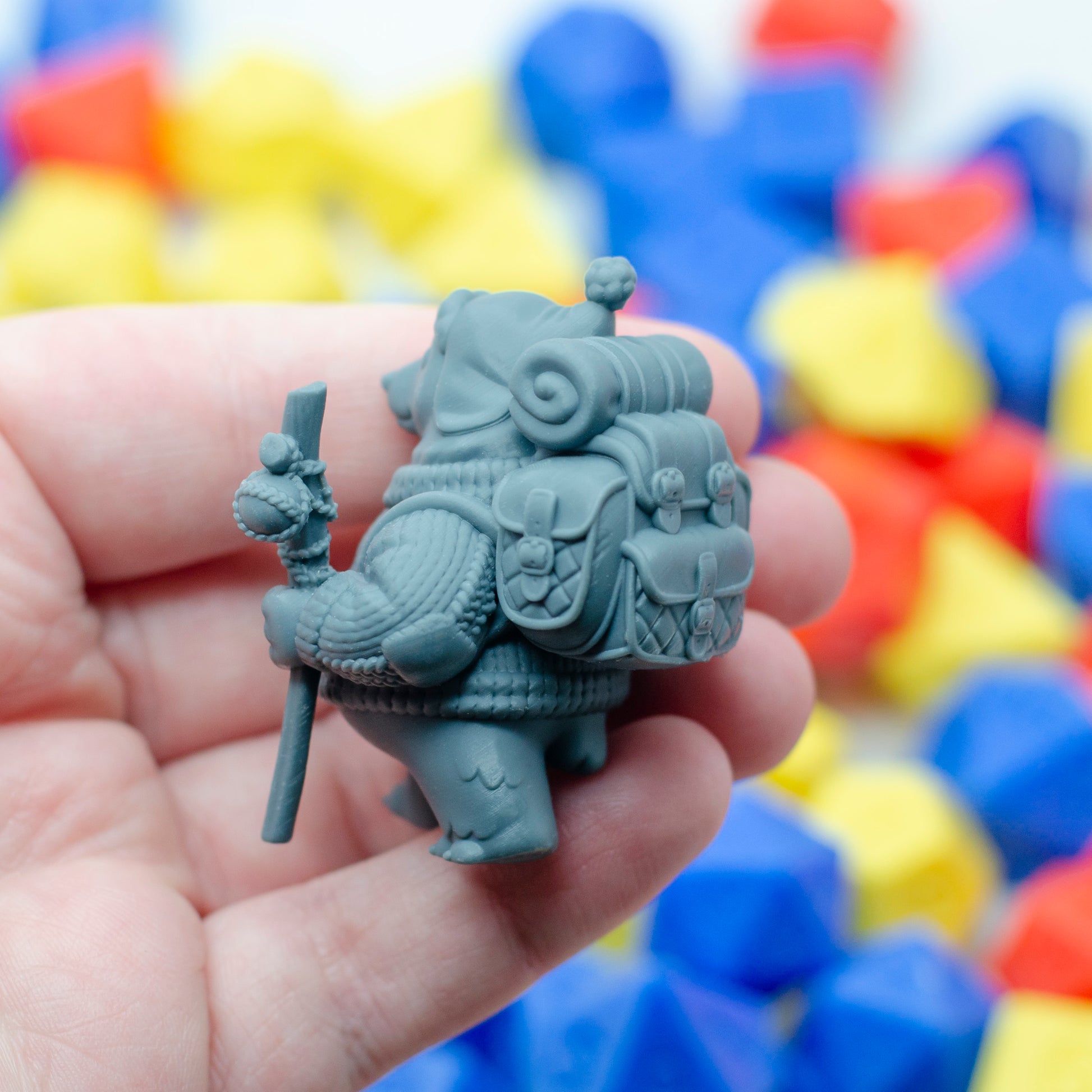 Gray 3d printed figurine of a traveling polar bear held in a hand with colorful blurred dice in the background. The backview shows the quilted details on the backpack and bedroll.