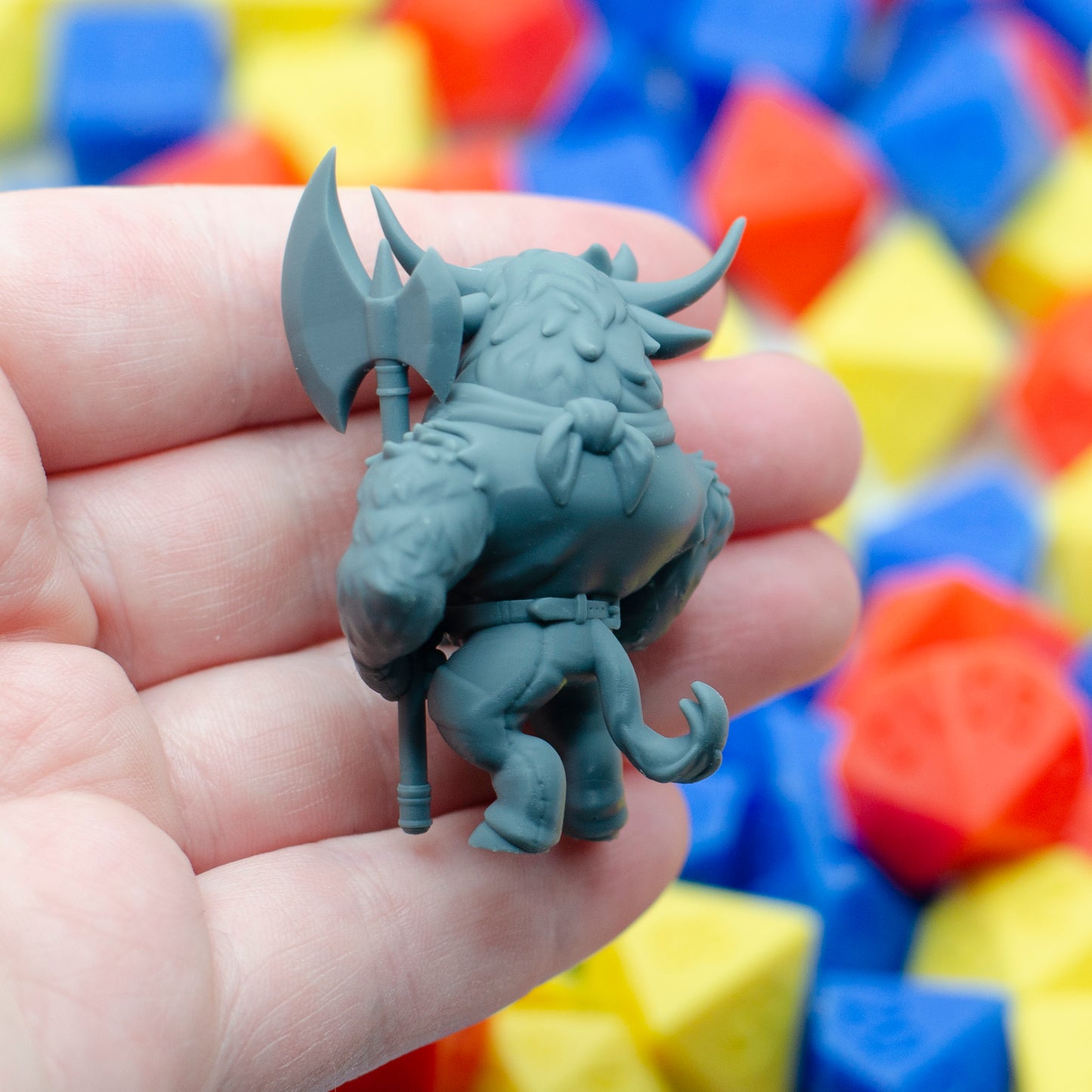 Gray 3d printed figurine of a minotaur, shown from the back and held in a hand with colorful dice in the background.