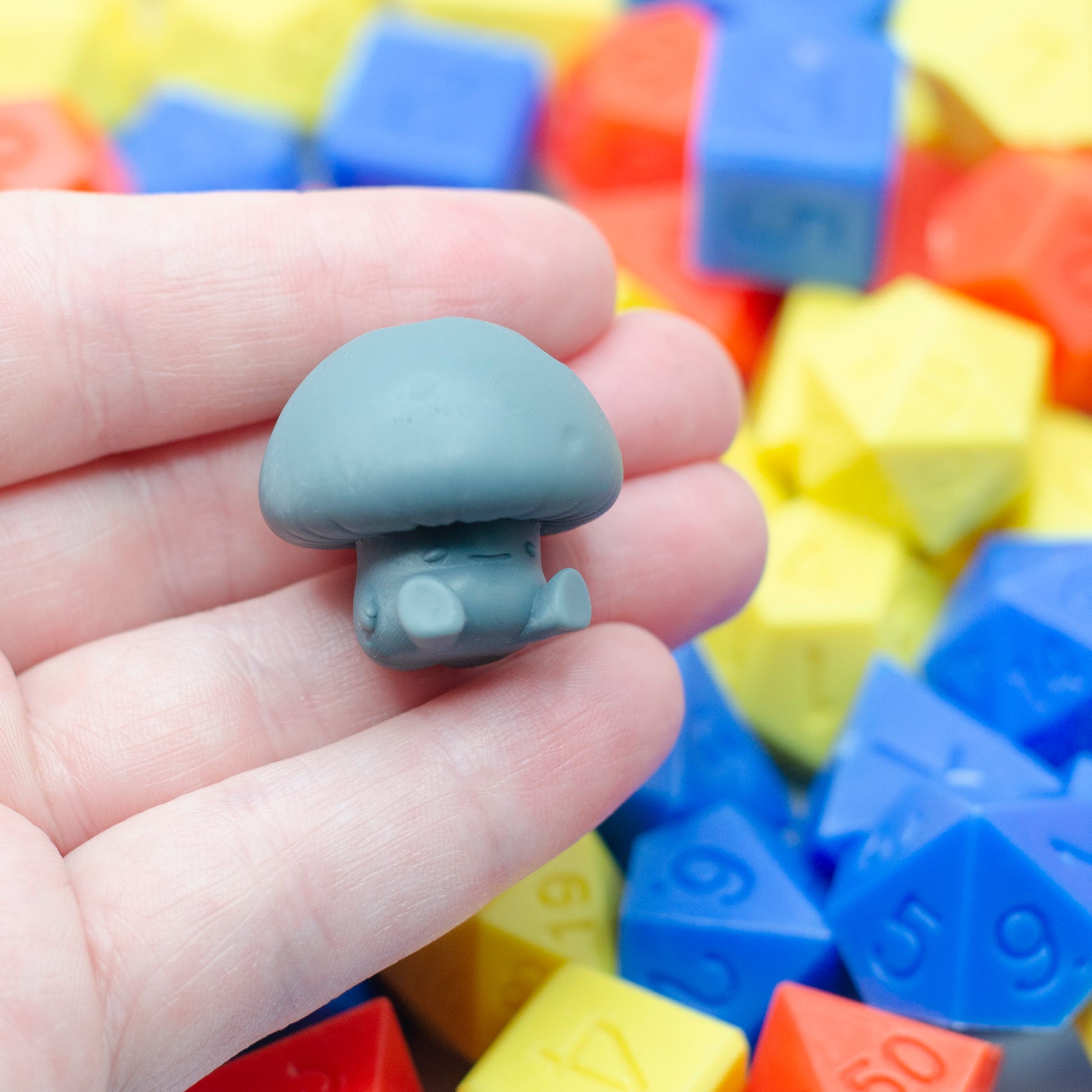 Gray 3d printed resin mushroom-shaped  hunched forward with a tired expression. The character is held in a hand, with colorful dice in the background.