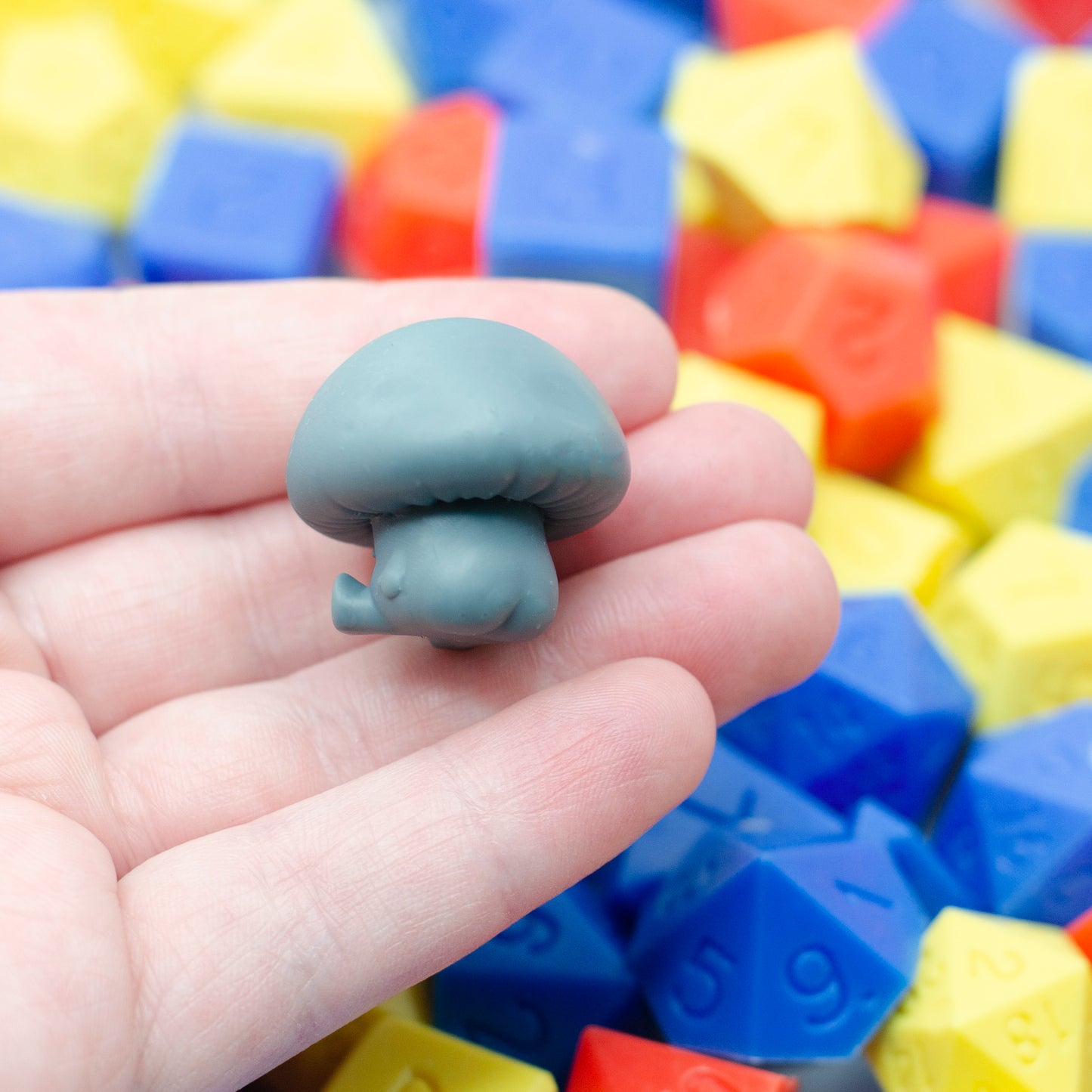 Gray 3d printed mushroom-shaped creature shown from the back, revealing its booty. It is held in a hand with colorful dice in the background.