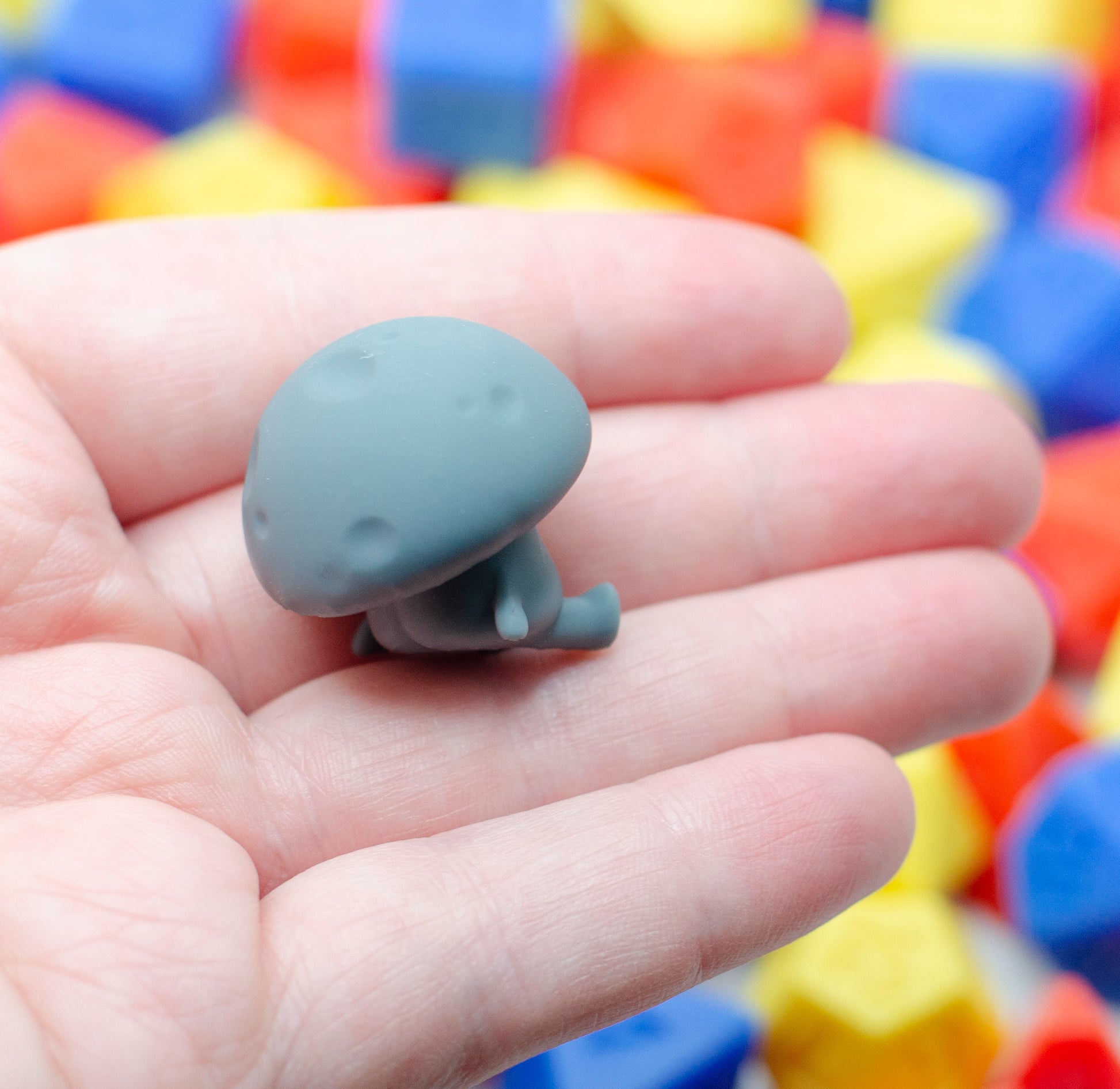 Small gray mushroom figurine leaning back with an exhausted expression. It is held in a hand with a colorful background